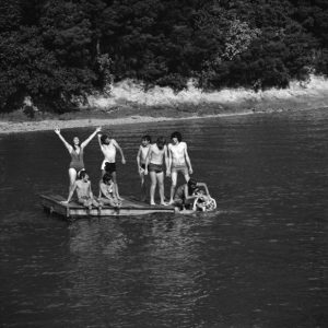 A group of people standing on a raft in the middle of a river. A young woman is raising her arms up to the sun, and the river looks warm and peaceful.